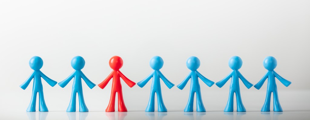 Row of seven plastic figurines holding hands, with six blue figures and one red figure standing in the center, symbolizing individuality or standing out from the crowd.