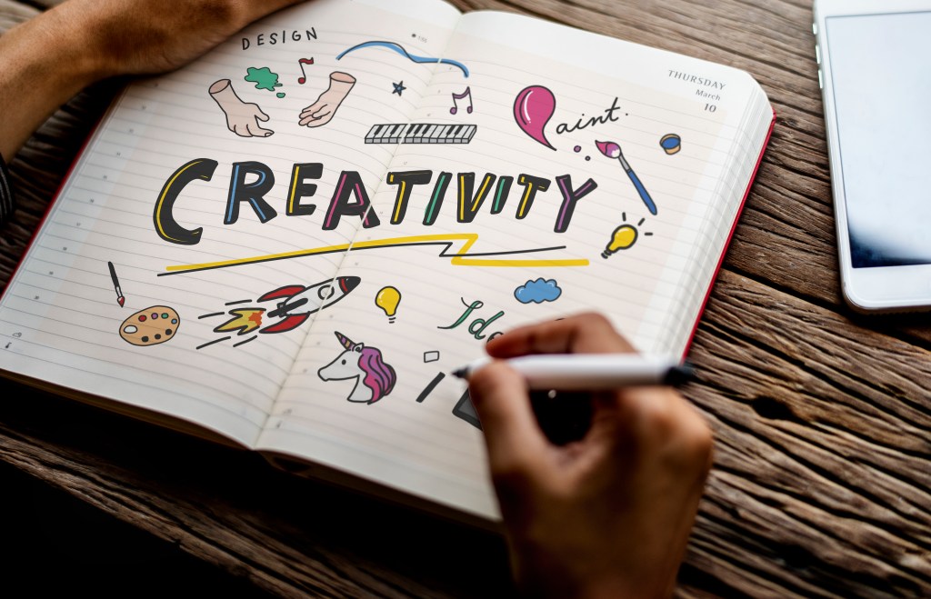 Person writing or drawing in an open notebook filled with colorful doodles and the large word 'CREATIVITY' in the center, surrounded by images such as a rocket, unicorn, light bulbs, paintbrush, and musical notes. The setting includes a wooden table and a smartphone placed nearby.