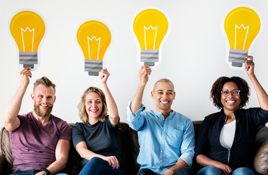 Four diverse people sitting on a couch, smiling and holding up large cutouts of light bulbs above their heads