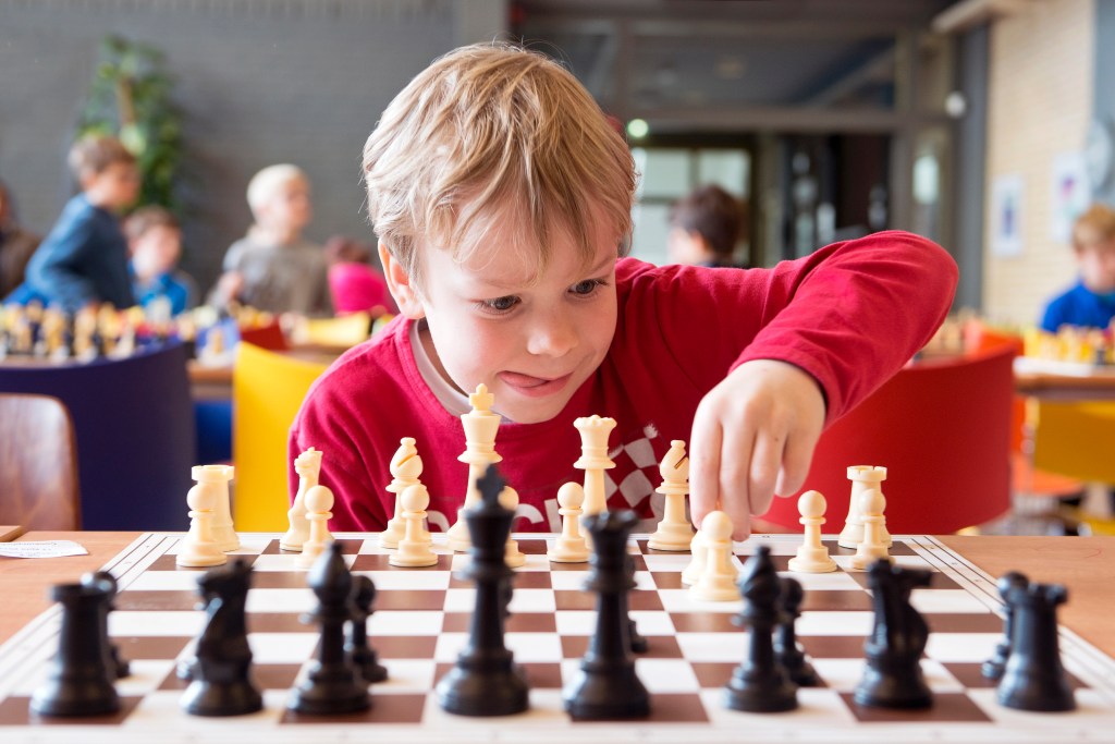 "A young child with blonde hair, wearing a red shirt, is intensely focused on a chessboard, making a move during a game. Other children can be seen in the blurred background, indicating a chess class or tournament setting."