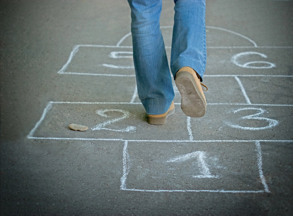 "A close-up of a person playing hopscotch on a chalk-drawn grid on the pavement. The person is mid-jump, with one foot in the air and the other on the ground, while a stone rests on the number 2 square."