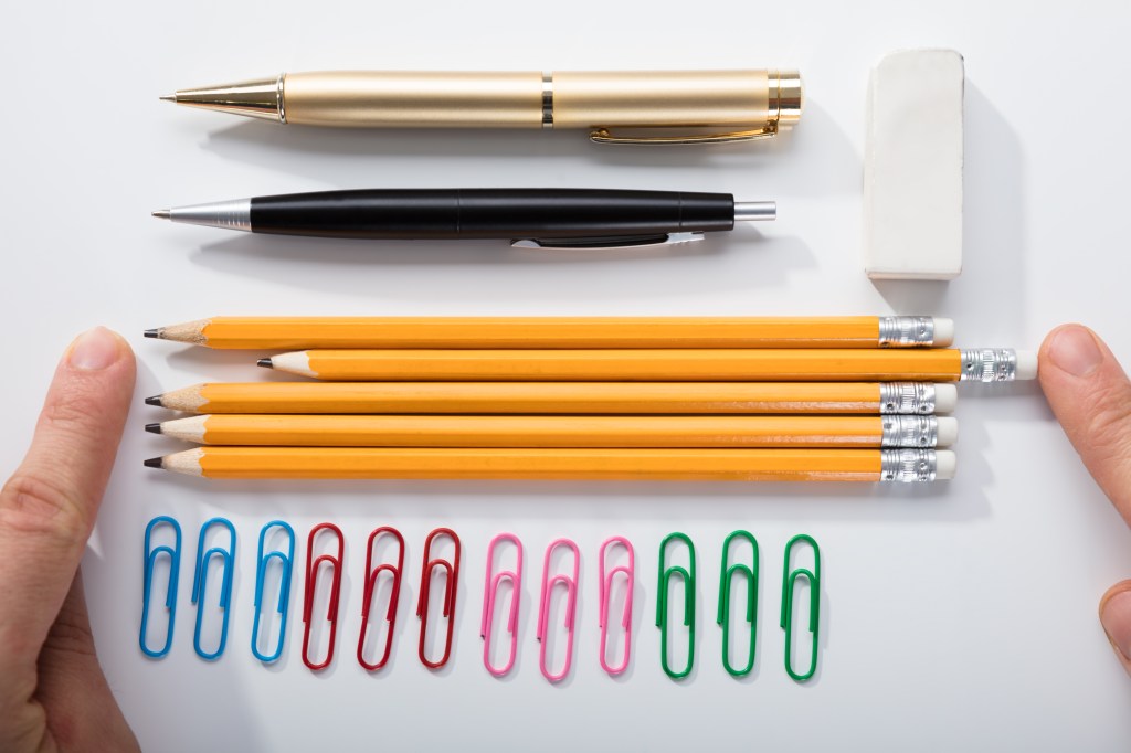 "Neatly arranged office supplies on a white surface, including two pens (one gold, one black), an eraser, six sharpened yellow pencils, and a row of colorful paper clips. Two fingers are seen on either side of the arrangement, pointing towards the items."