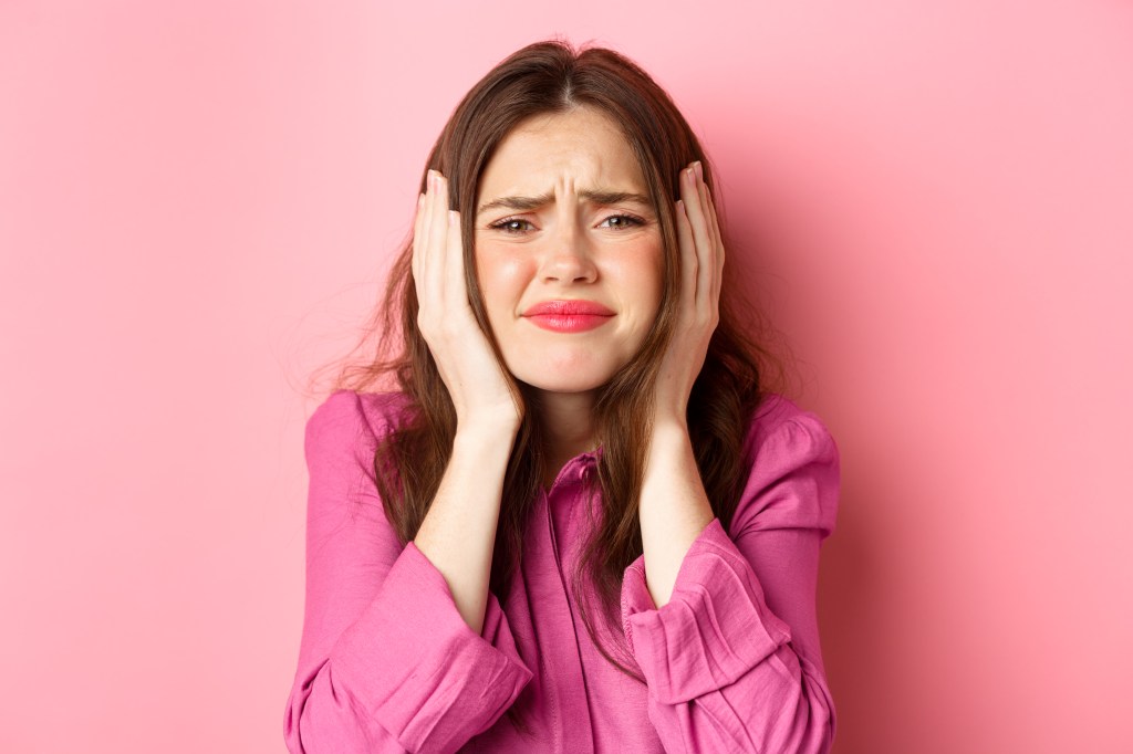"A young woman with long brown hair, wearing a pink shirt, holds her hands over her ears with a concerned or distressed expression on her face. The background is solid pink, matching her shirt."