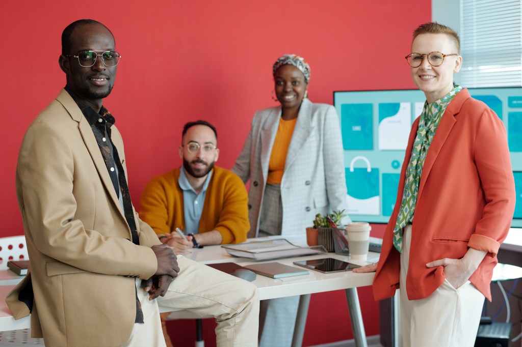 Group of diverse colleagues in contemporary office. Four professionals posing around table with digital screen in background, fostering collaborative and creative environment