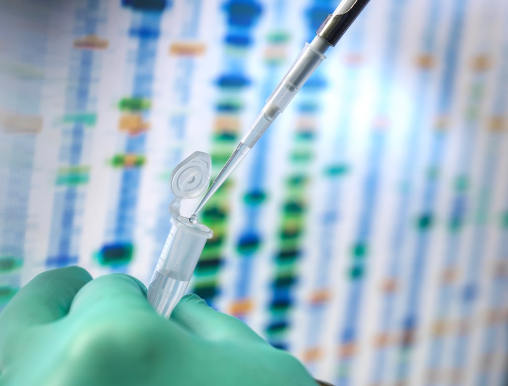 This image shows a close-up of a laboratory setting, where a gloved hand holds a small vial while a pipette dispenses liquid into it. In the background, there is a blurred representation of a DNA sequence or genetic data, highlighting a focus on genetic research, biotechnology, or molecular biology experiments.
