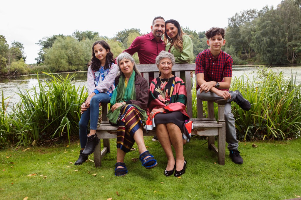 This image features a multi-generational family posing together outdoors by a lake. Two elderly women are seated on a wooden bench, surrounded by younger family members, including a girl, a boy, and two adults standing behind the bench. The scene is peaceful, with greenery and trees in the background.