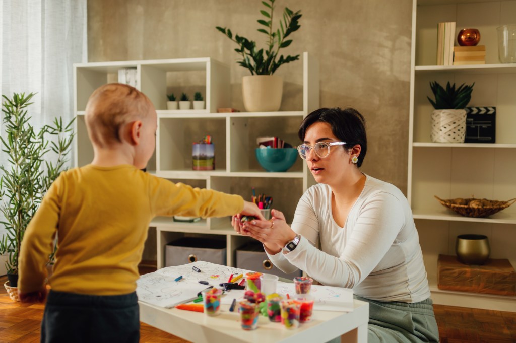 This image shows a woman and a young child engaged in an activity at a small table in a cozy, well-decorated room. The woman, wearing glasses and a light top, is seated, accepting an object or toy from the child, who is standing and wearing a yellow shirt. The table is covered with art supplies, including colorful crayons and drawings, suggesting they are doing arts and crafts together. The room has a calm atmosphere with plants and shelves in the background, making it a warm, educational environment.